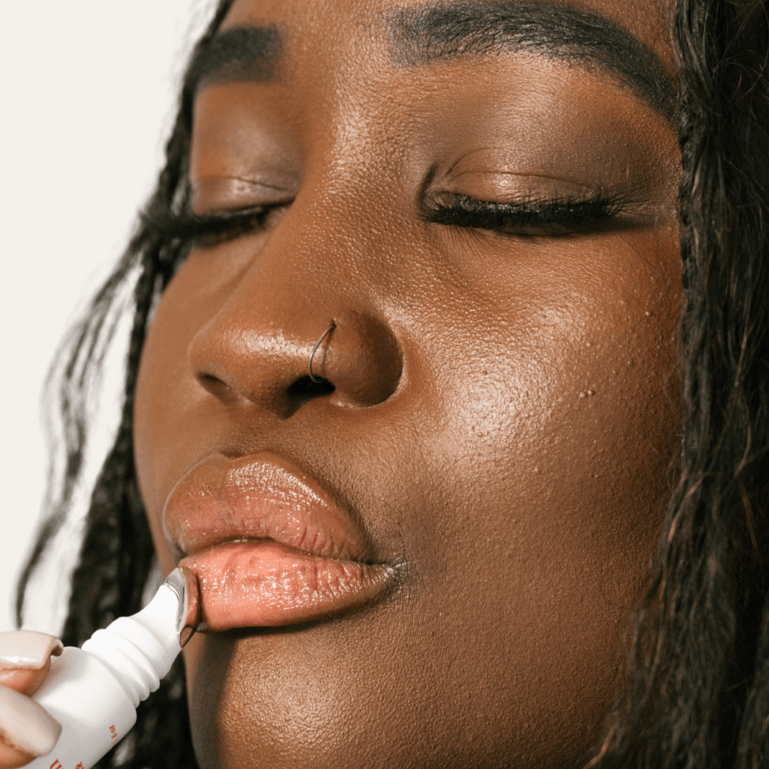 A woman applying a The Balm, a nourishing and healing lip balm to their lips with a white background