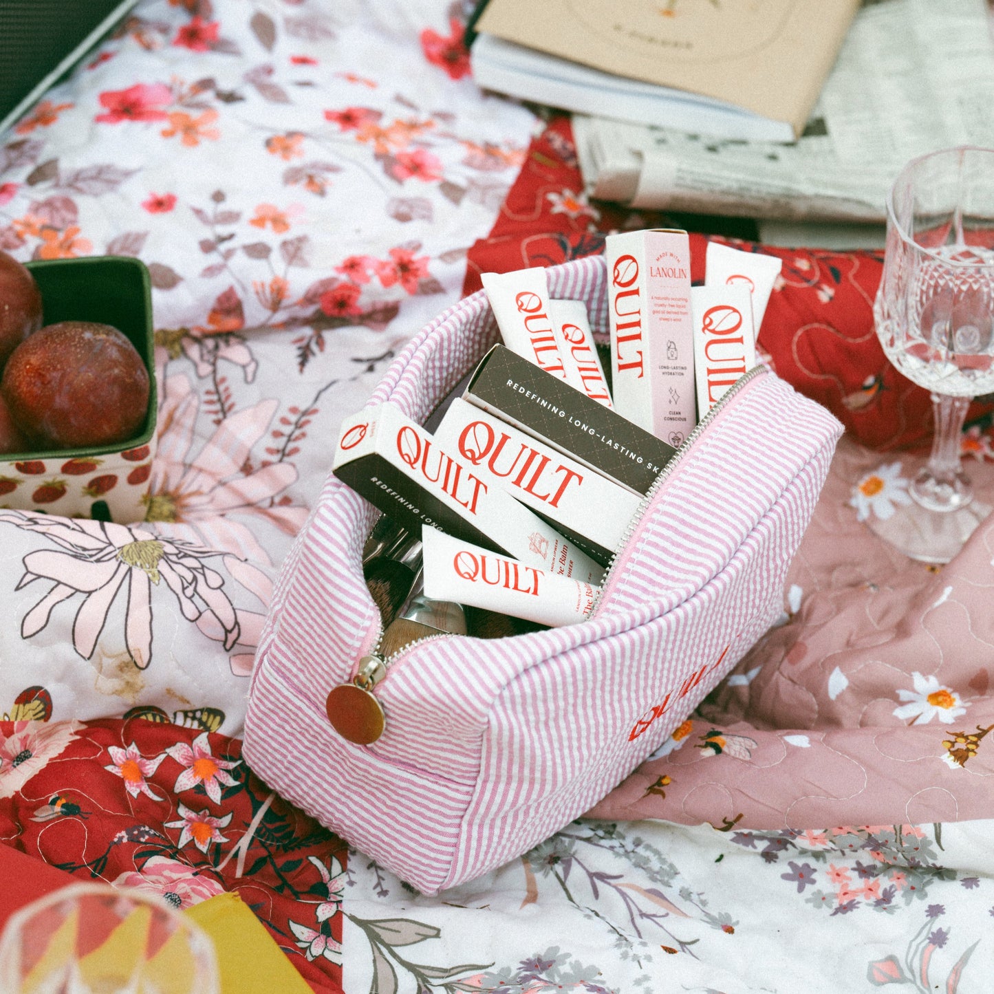 QUILT's The Balm in a pink makeup bag on a floral tablecloth with books, glasses and fruits.