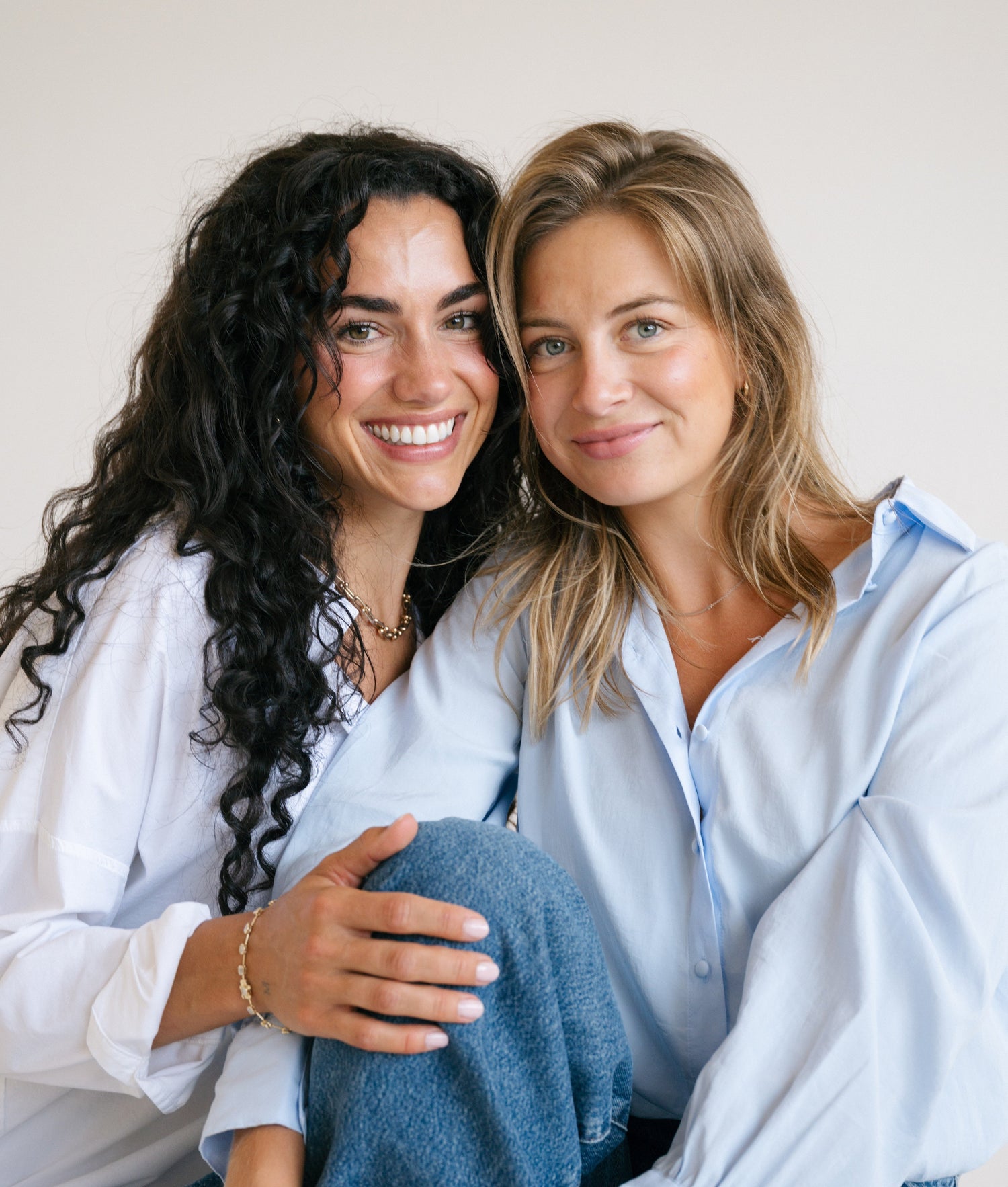Two women sitting together wearing light blue shirts against a plain background